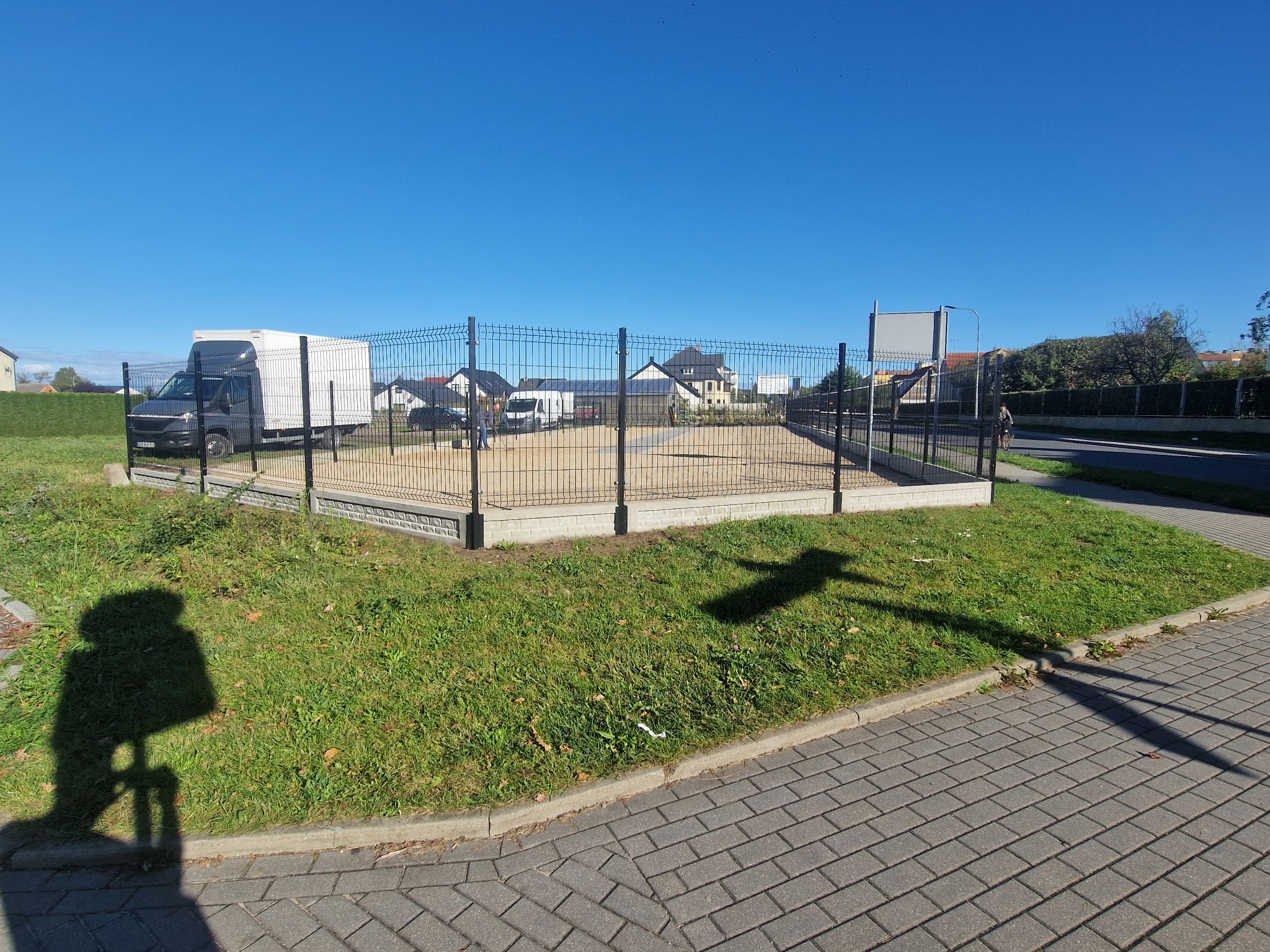 Ciemnoszara siatka ogrodzeniowa na betonowej podmurówce, otaczająca teren z piaskiem; w tle zaparkowany biały samochód dostawczy i zabudowa jednorodzinna w słoneczny dzień.