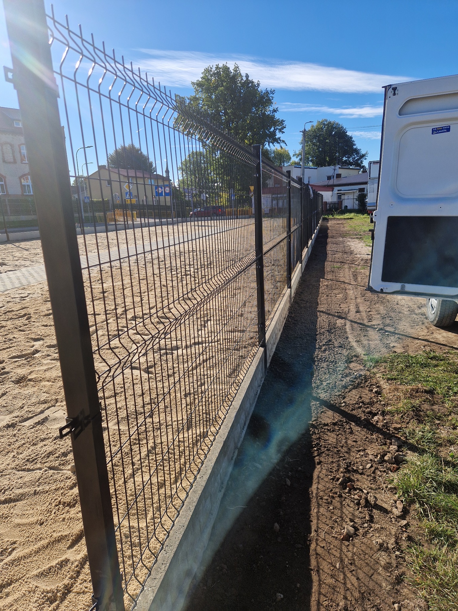 Nowo zainstalowane ogrodzenie panelowe z podmurówką betonową, widoczne z perspektywy gruntu, z piaskiem po jednej stronie i świeżo przekopaną ziemią po drugiej, obok zaparkowany biały bus.