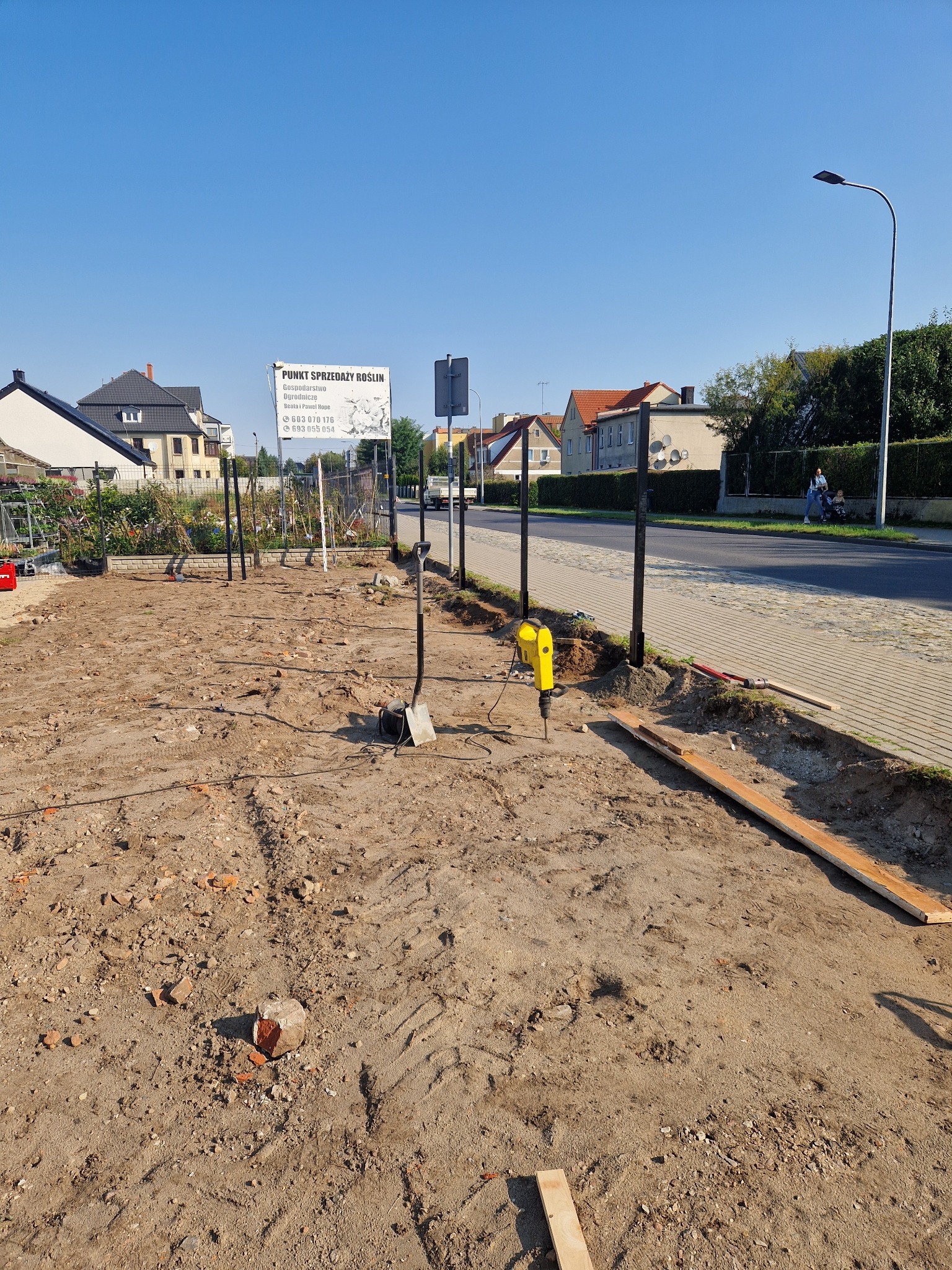 Przygotowany teren pod montaż ogrodzenia z czarnymi słupkami; widoczny żółty młot udarowy wbijający słupek, ziemia z gruzem, w tle chodnik i zabudowania.