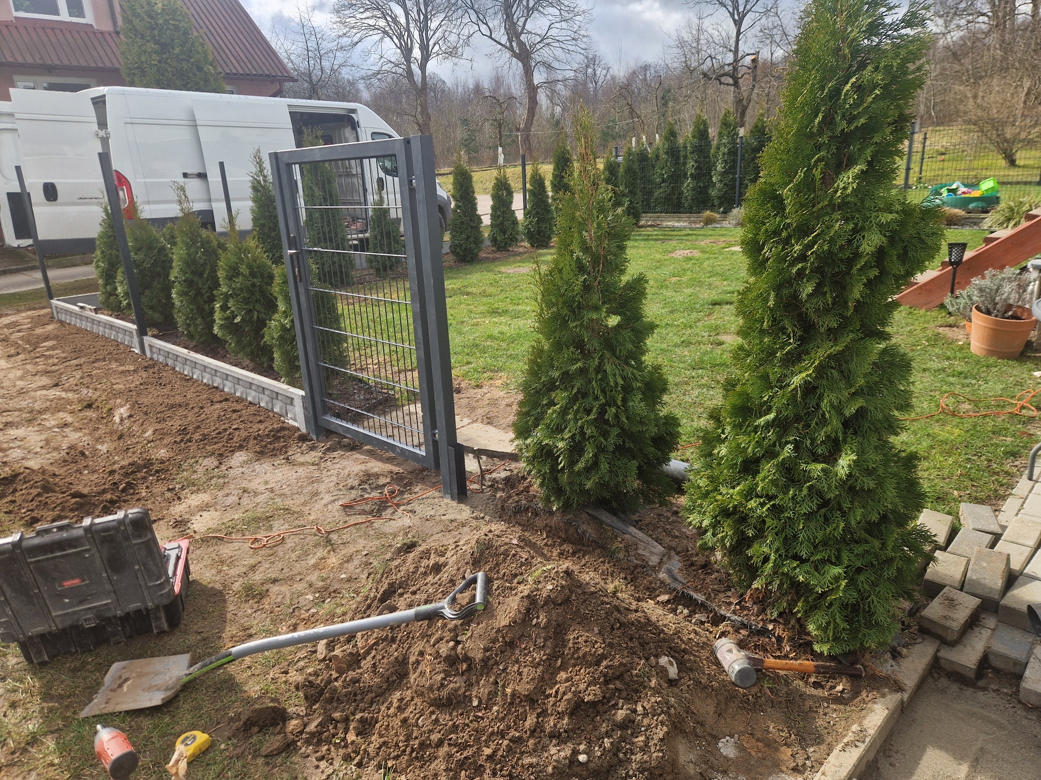 Montaż ogrodzenia panelowego z siatki w trakcie prac ziemnych, widoczna brama, narzędzia, sadzonki tui i dostawczy samochód w tle.