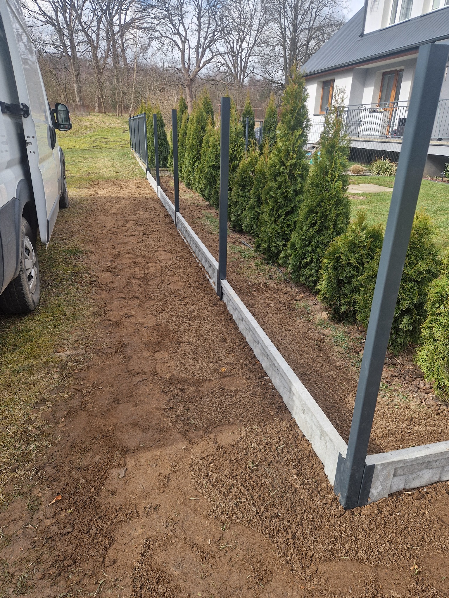 Montaż ogrodzenia panelowego z podmurówką betonową, widoczne słupki i fragment siatki, obok zaparkowany biały bus.