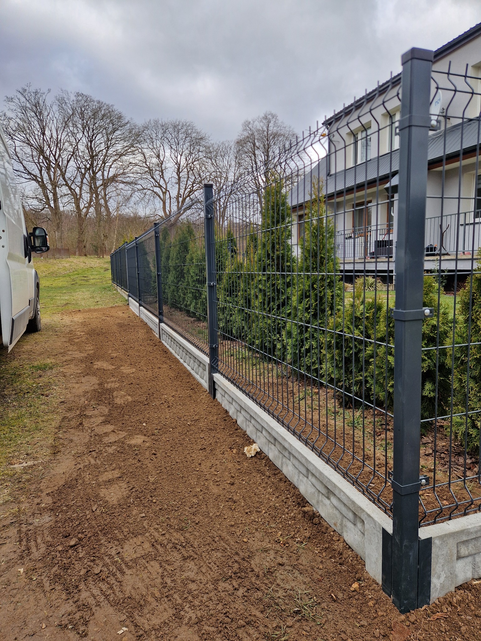 Nowoczesne ogrodzenie panelowe z podmurówką betonową, widoczne z boku, obok zaparkowany biały samochód dostawczy, w tle dom i drzewa.