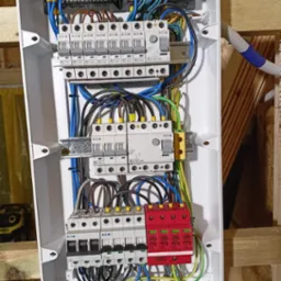 Otwarta skrzynka rozdzielcza z bezpiecznikami EATON i ogranicznikami przepięć ELS, widoczne kable w kolorach niebieskim, brązowym, żółto-zielonym i czarnym, zamontowana na drewnianej konstrukcji.