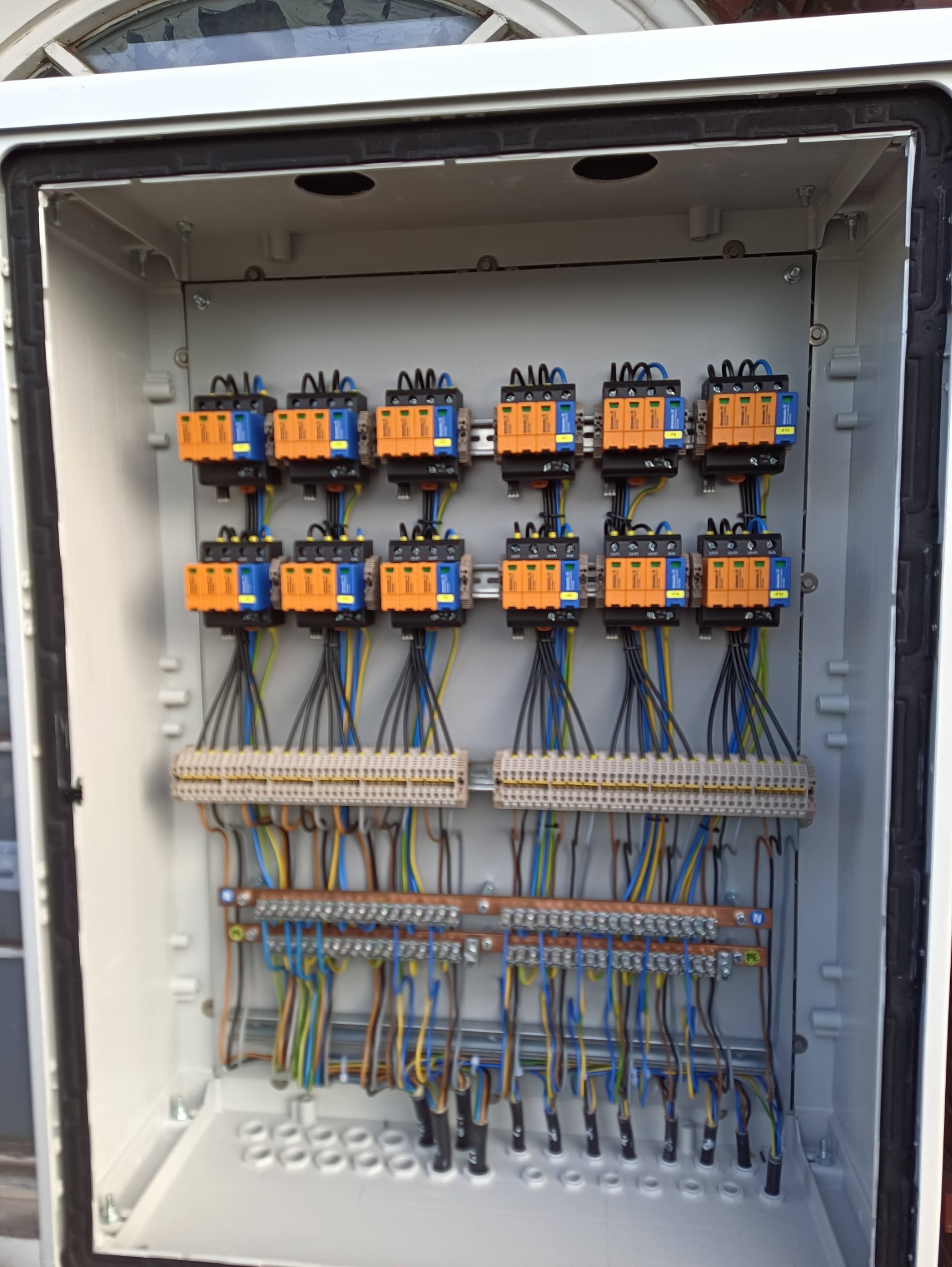 Wewnętrzny widok skrzynki elektrycznej z zamontowanymi ogranicznikami przepięć w kolorze pomarańczowo-niebieskim, listwami zaciskowymi i okablowaniem w różnych kolorach.