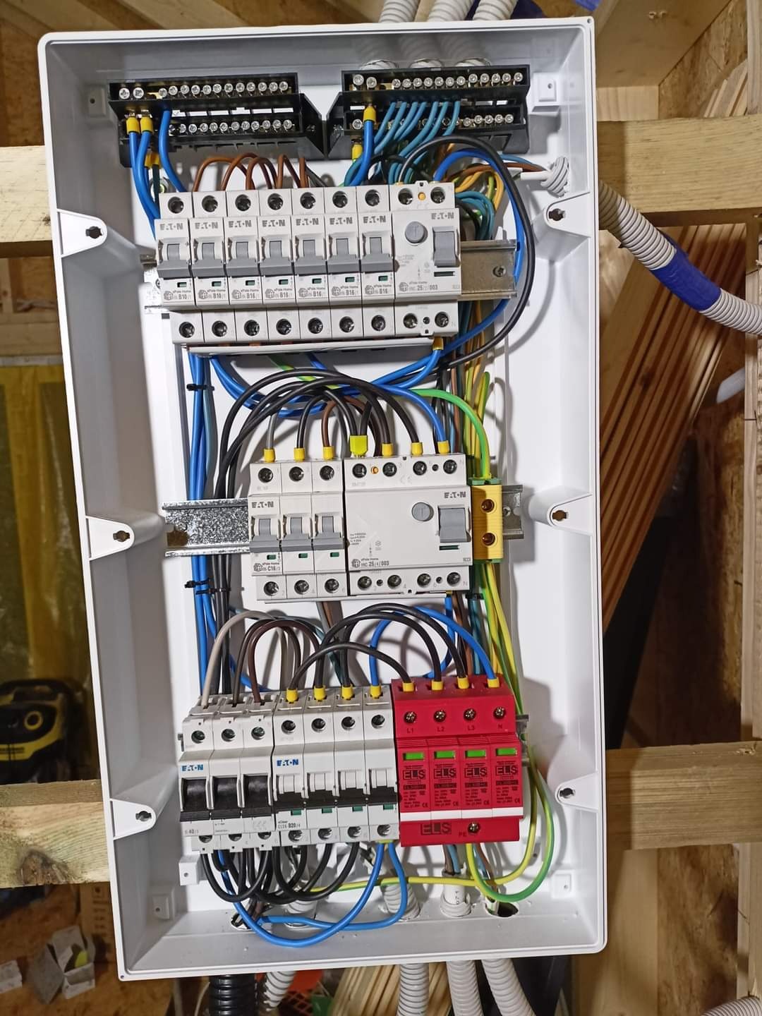 Otwarta skrzynka rozdzielcza z bezpiecznikami EATON i ogranicznikami przepięć ELS, widoczne kable w kolorach niebieskim, brązowym, żółto-zielonym i czarnym, zamontowana na drewnianej konstrukcji.