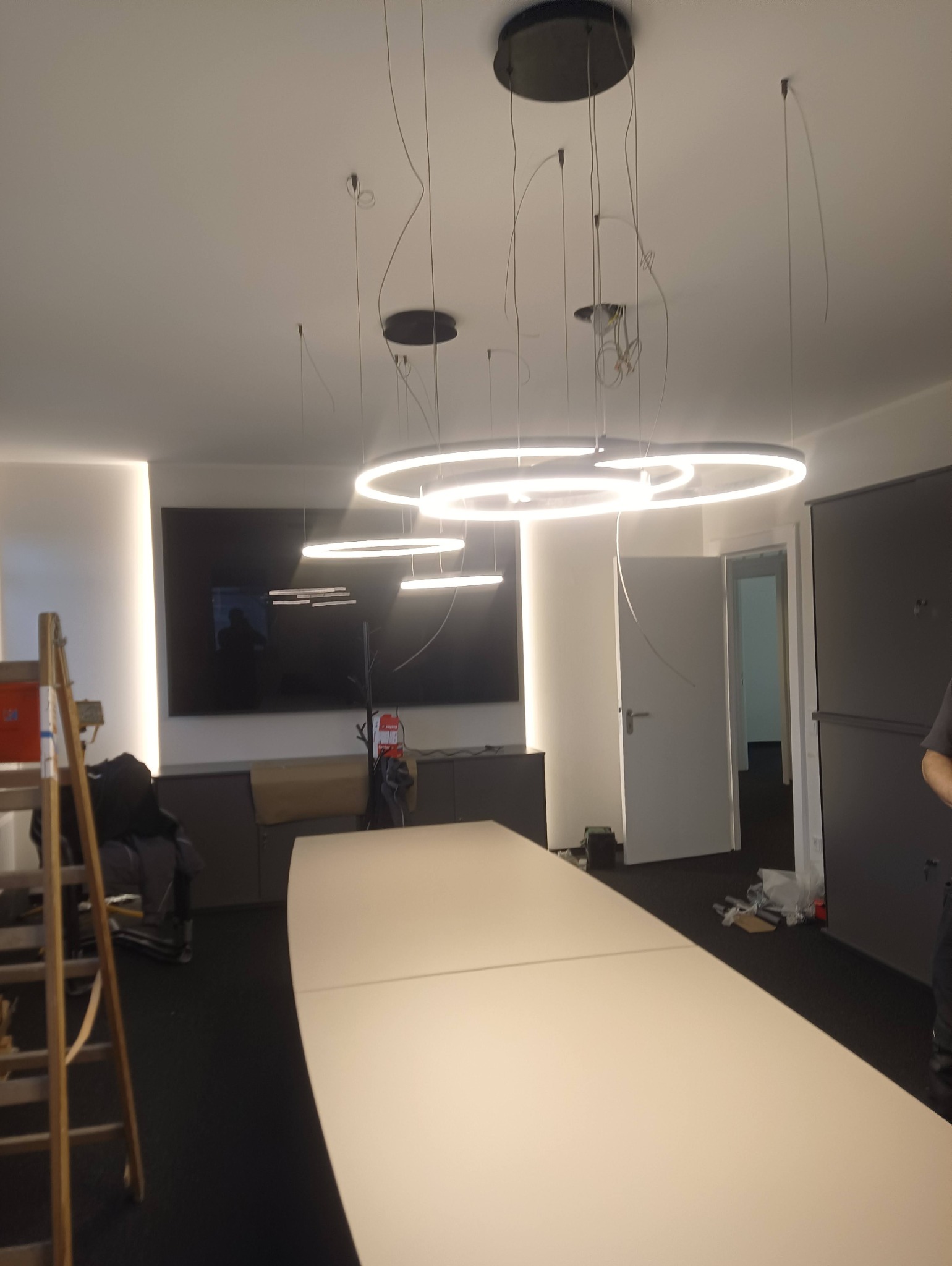 Montaż nowoczesnych, okrągłych lamp LED w biurze, widoczne przewody i drabina malarska, sufit z czarnymi mocowaniami.