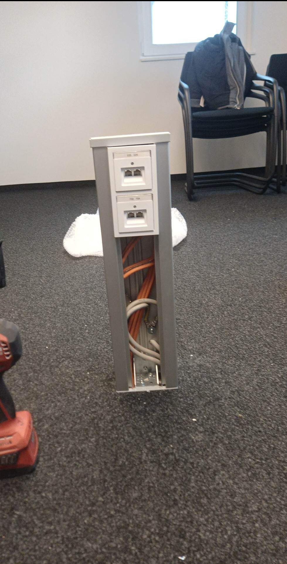 Szara kolumna z gniazdami sieciowymi i otwartym kanałem kablowym, wewnątrz widoczne pomarańczowe i białe przewody, w tle biurowe krzesła i okno, na pierwszym planie fragment wiertarki akumulatorowej.