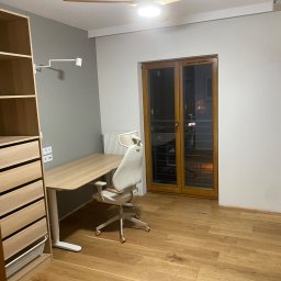 DWELLTOWER - Pokój z drewnianą podłogą, biurkiem z regulowaną wysokością, ergonomicznym krzesłem obrotowym i szafą z jasnego drewna, oświetlony lampą sufitową i bocznym kinkietem.