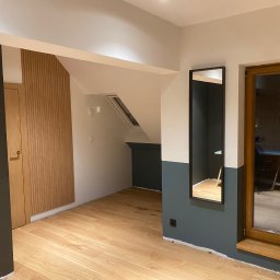 DWELLTOWER - Wnętrze po remoncie: drewniana podłoga, ściany pomalowane w dwóch kolorach (biały i ciemny szary), drewniane drzwi z pionowymi panelami, duże okno w drewnianej ramie i lustro w czarnej ramie.