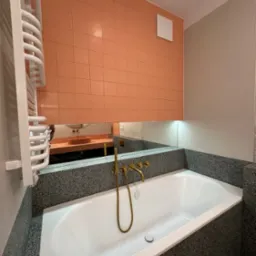 Wykończona łazienka z białą wanną obudowaną szarym lastryko, złotą armaturą i pomarańczowymi płytkami na ścianie, widoczna również w lustrze toaletka z umywalką.