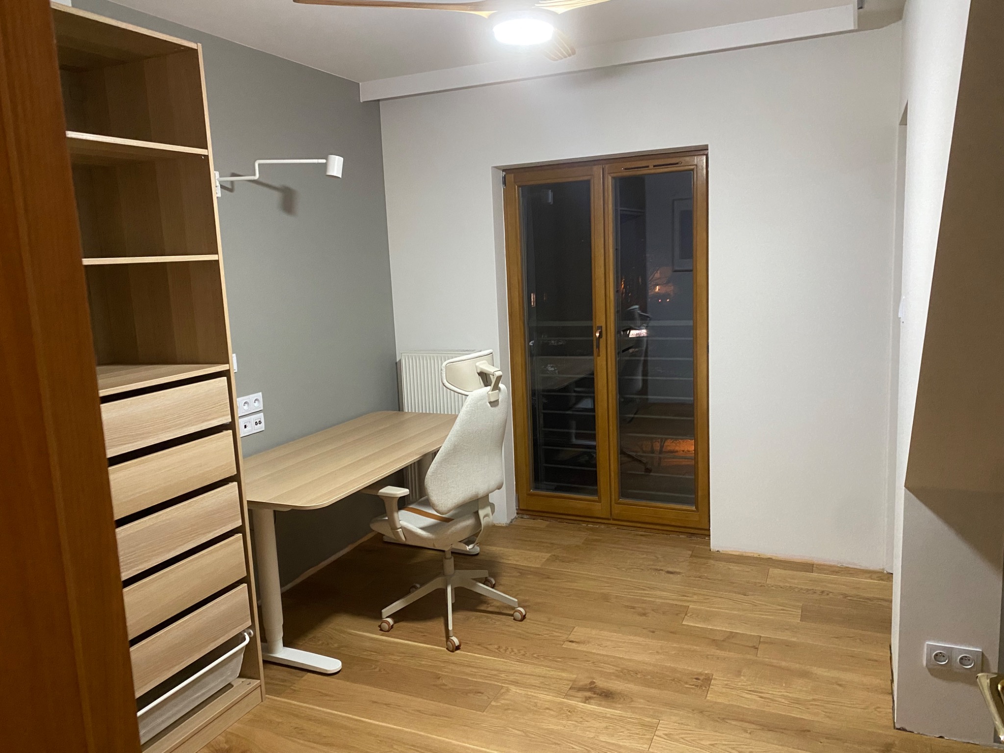 Pokój z drewnianą podłogą, biurkiem z regulowaną wysokością, ergonomicznym krzesłem obrotowym i szafą z jasnego drewna, oświetlony lampą sufitową i bocznym kinkietem.