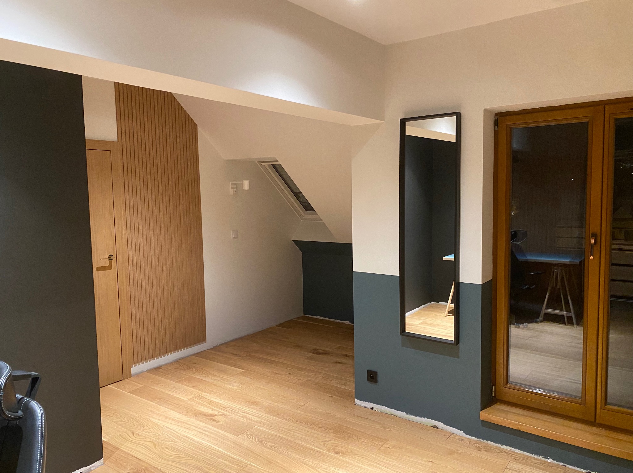 Wnętrze po remoncie: drewniana podłoga, ściany pomalowane w dwóch kolorach (biały i ciemny szary), drewniane drzwi z pionowymi panelami, duże okno w drewnianej ramie i lustro w czarnej ramie.