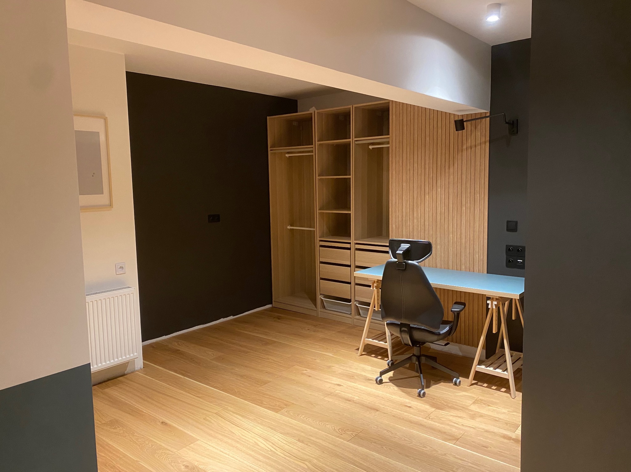 Pokój z drewnianą podłogą, szafą w zabudowie z drewnianymi lamelami, biurkiem na drewnianych nogach i czarną ścianą, oświetlony punktowym światłem.