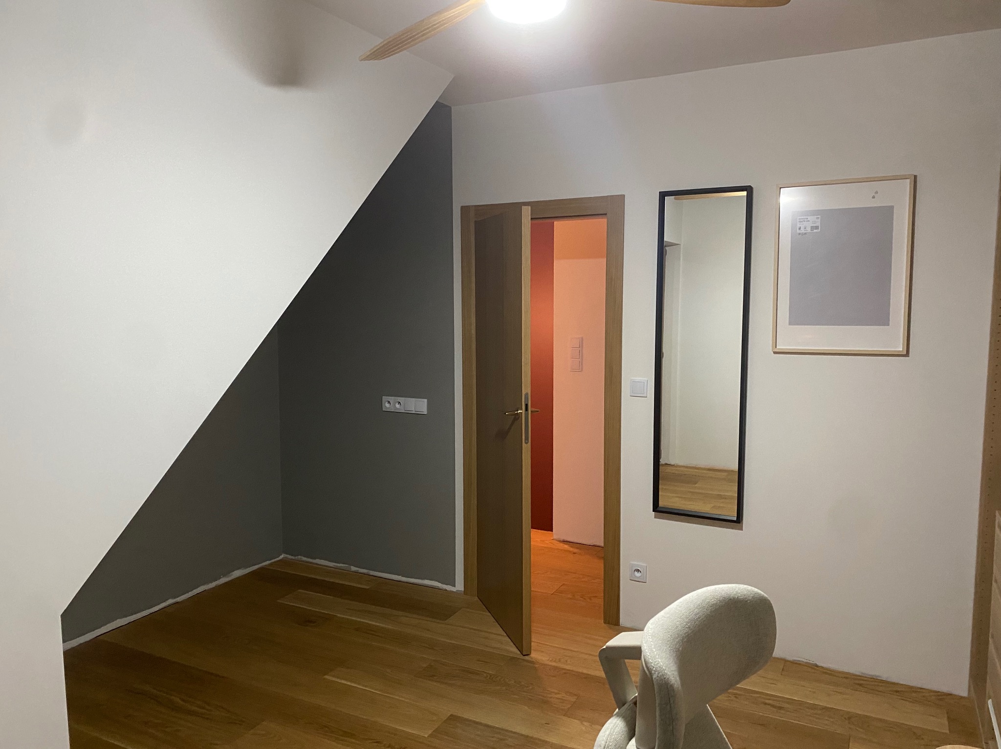 Pokój na poddaszu po remoncie: pomalowane ściany w kolorze białym i szarym, drewniana podłoga, otwarte drewniane drzwi, lustro w czarnej ramie i obraz na ścianie.