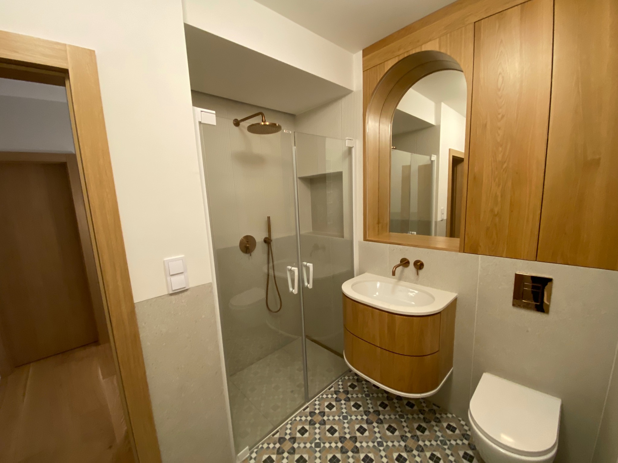 Nowoczesna łazienka z kabiną prysznicową, umywalką na szafce z drewnianymi frontami, toaletą i wzorzystą podłogą. Armatura w kolorze miedzi, a nad umywalką szafka z lustrem w kształcie łuku.