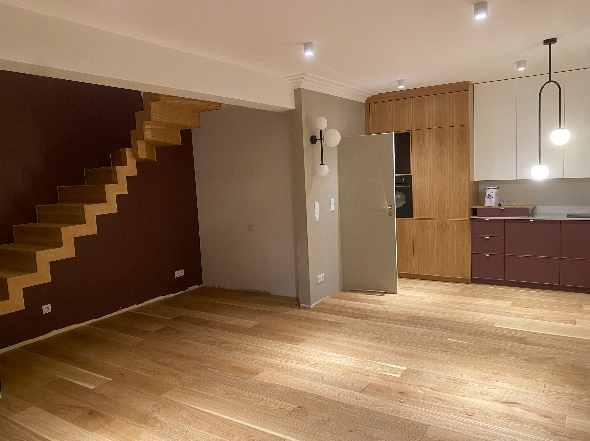 Nowoczesne wnętrze po remoncie: drewniane schody bez balustrady na tle bordowej ściany, jasna drewniana podłoga, otwarta kuchnia z drewnianymi i białymi szafkami oraz bordowymi dolnymi szafkami...