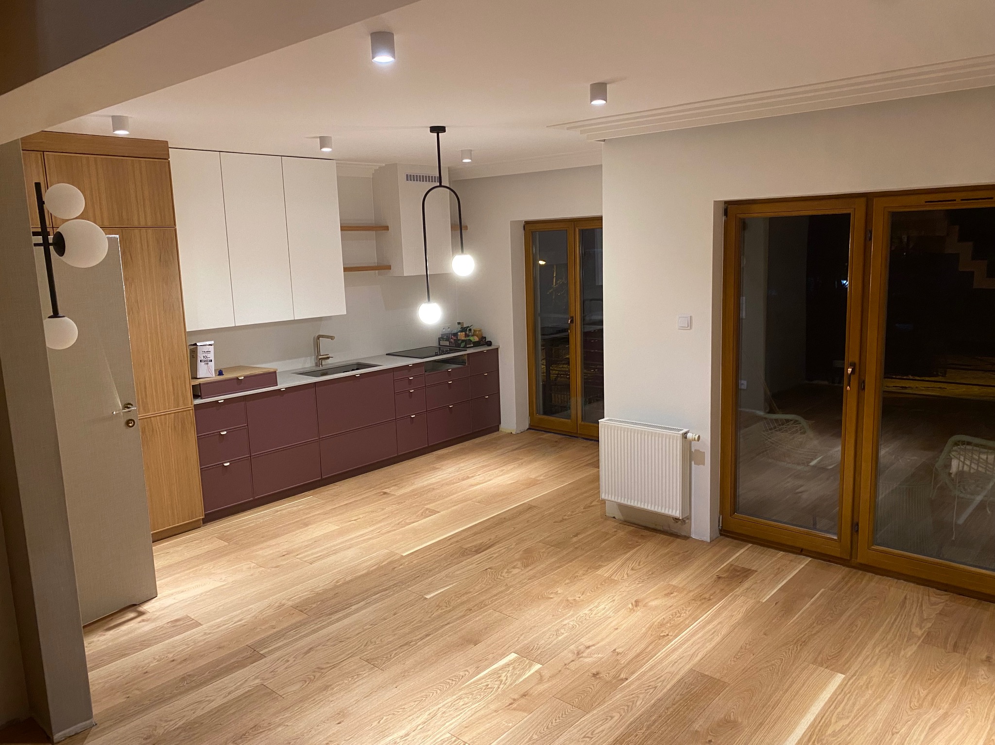 Nowoczesna kuchnia z drewnianą podłogą, bordowymi szafkami dolnymi i białymi górnymi, oświetlona wiszącą lampą z dwoma kulami, widok na drewniane okna tarasowe i grzejnik.