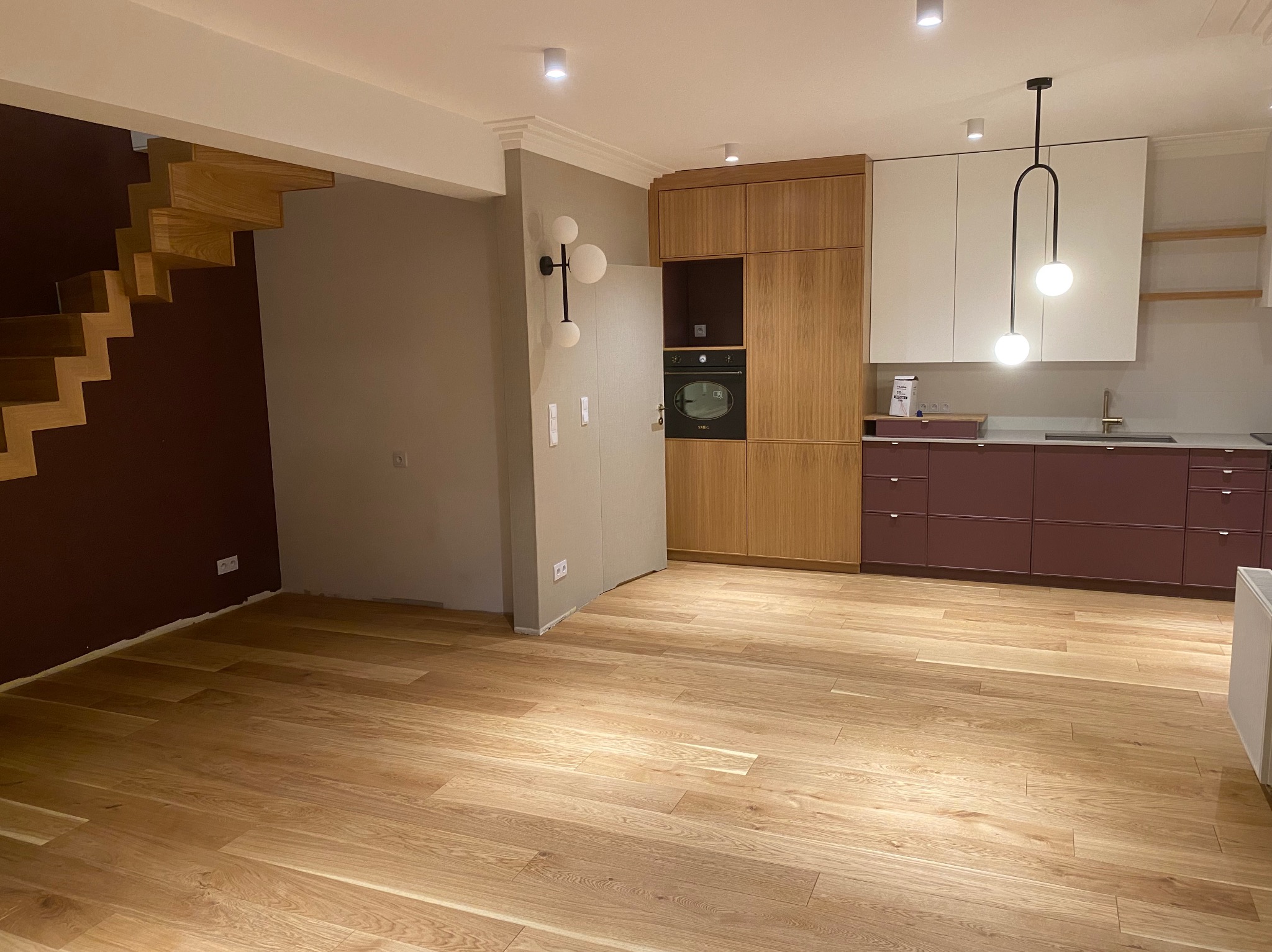 Nowoczesna kuchnia w Gdyni z drewnianymi schodami, zabudową kuchenną z elementami w kolorze burgundowym i złotą armaturą, oświetlona designerską lampą.