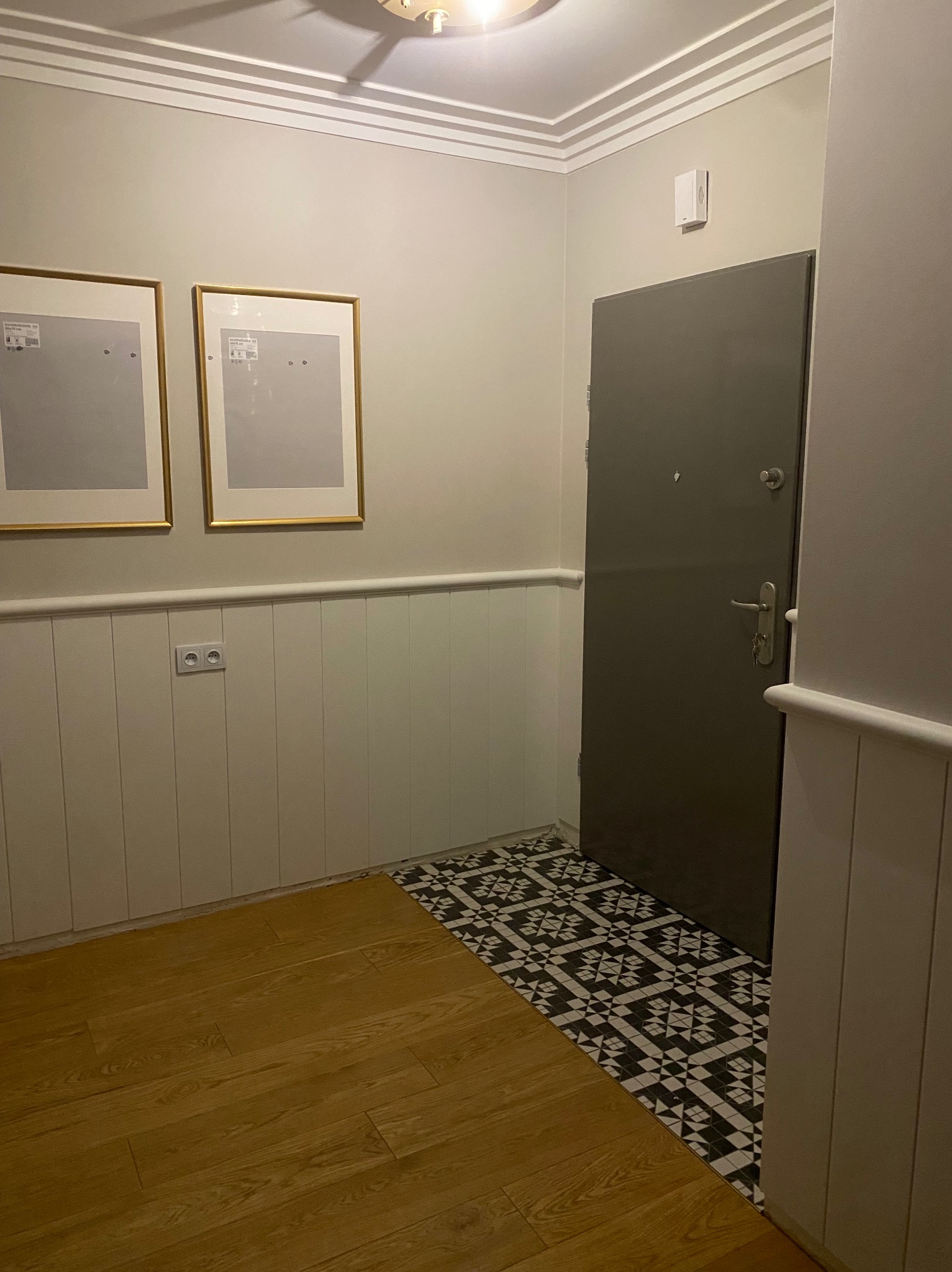 Korytarz po remoncie z panelami ściennymi, nową podłogą z płytek w geometryczny wzór i drewnianymi panelami, szara metalowa drzwi i dwoma pustymi ramkami na ścianie.