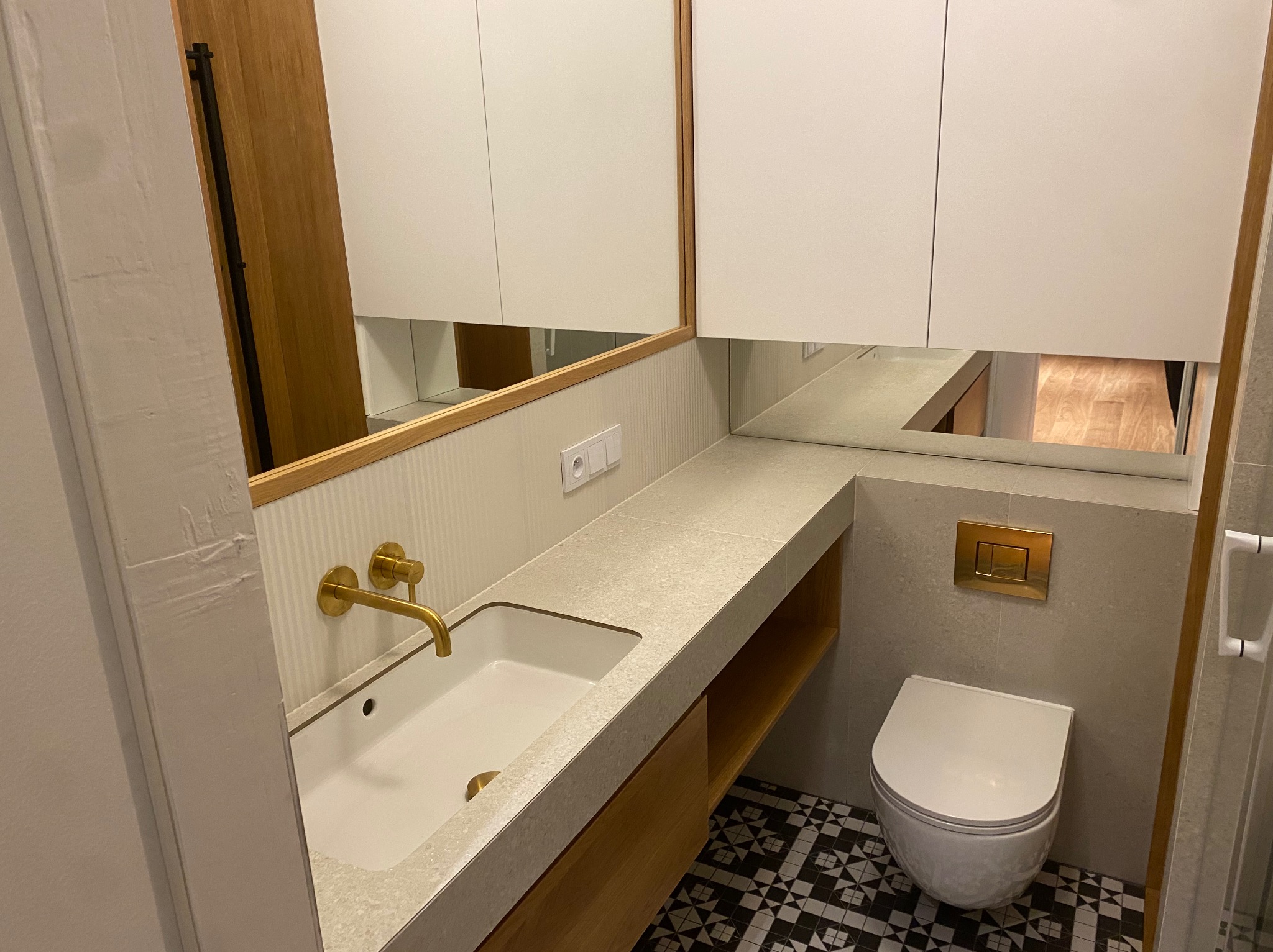 Nowoczesna łazienka z wiszącą toaletą, umywalką nablatową z baterią w kolorze starego złota, szafkami wiszącymi i podłogą w czarno-białą szachownicę.