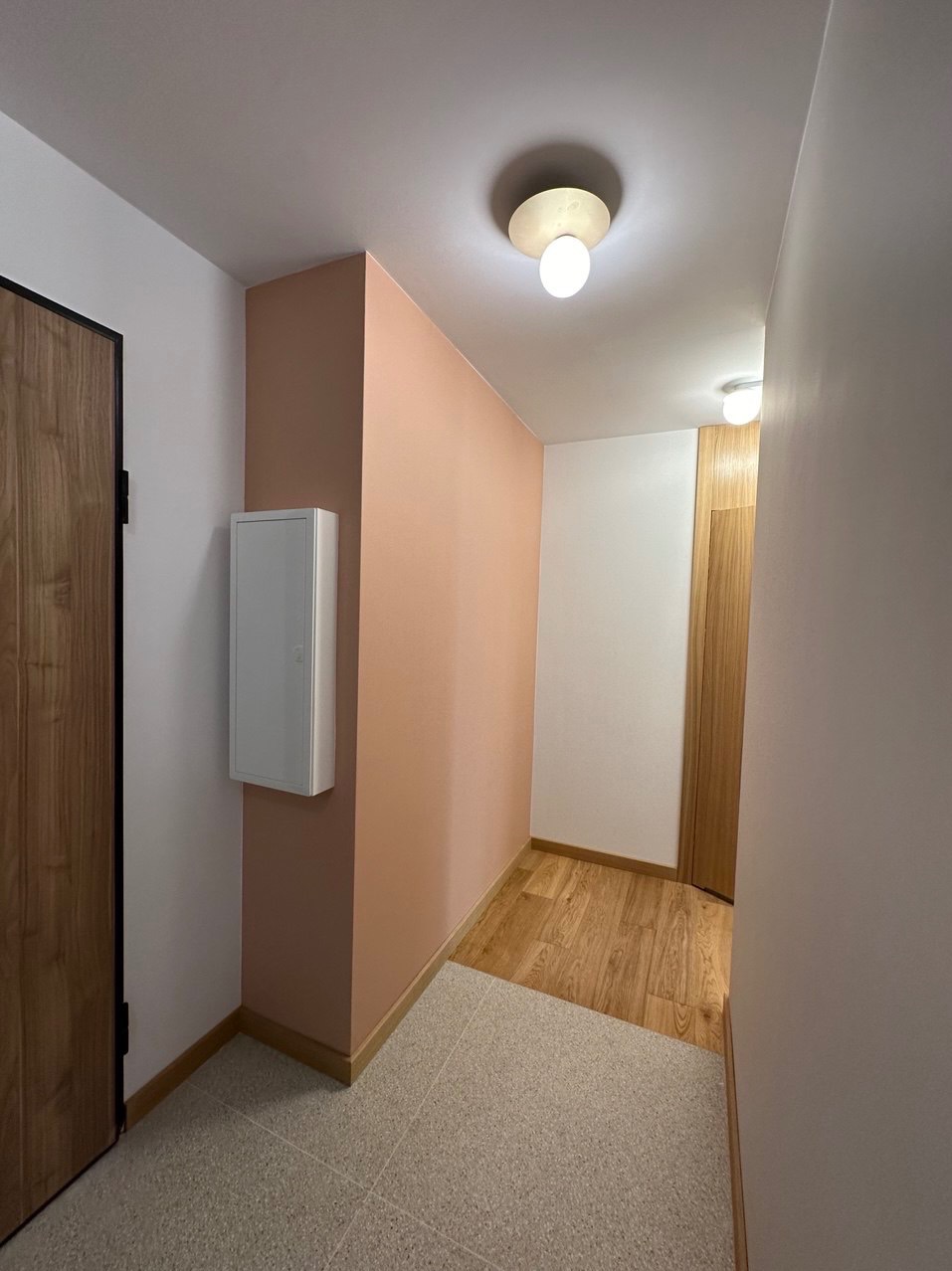 Korytarz po odświeżeniu: ściana pomalowana na brzoskwiniowy kolor, kontrastująca z białym sufitem i szafką elektryczną; podłoga wyłożona płytkami imitującymi lastryko, przechodząca w panelową...