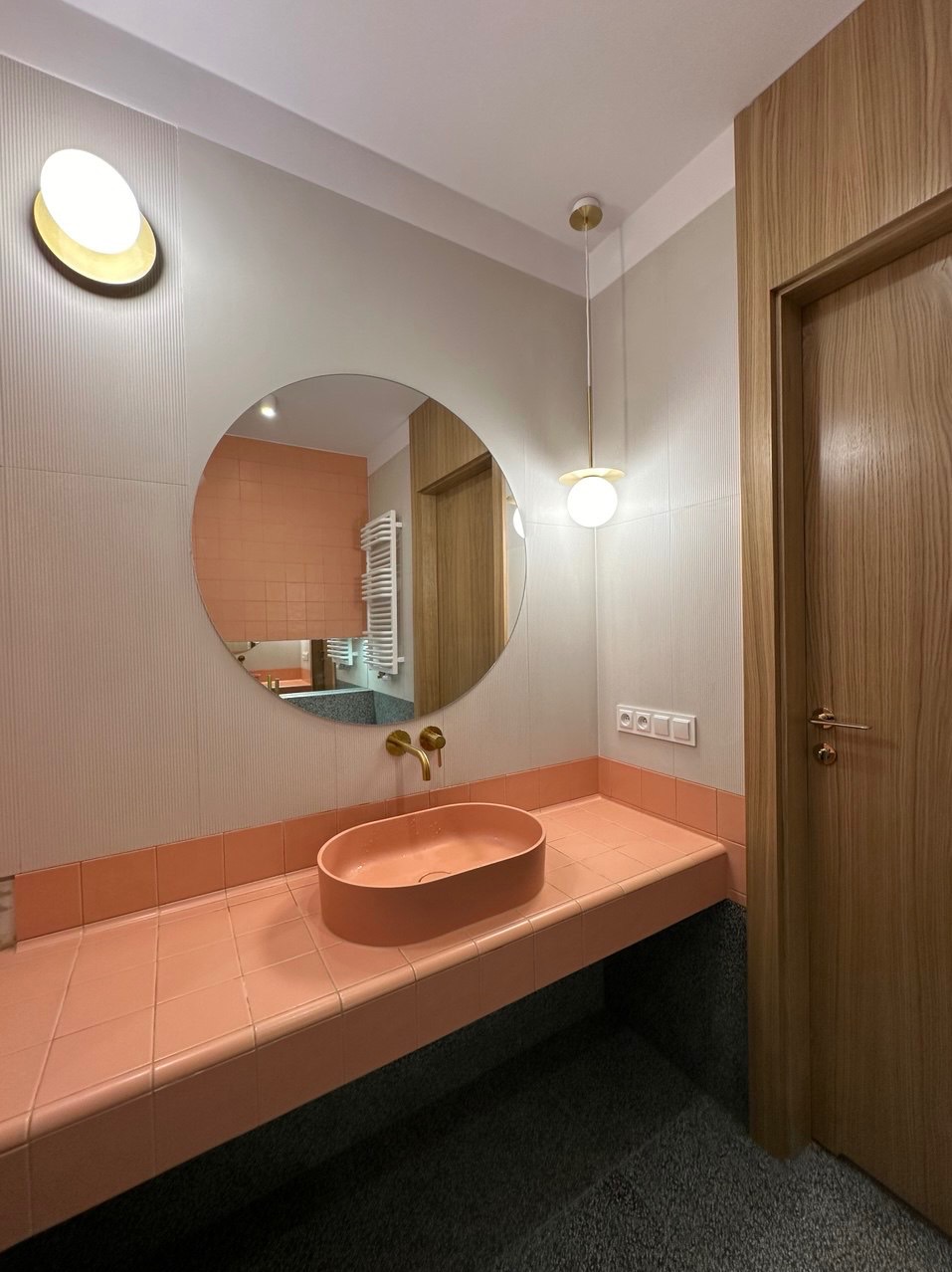 Nowoczesna łazienka z owalną umywalką w kolorze łososiowym, złotą armaturą i okrągłym lustrem, na tle ściany w pionowe prążki i drewnianych drzwi.