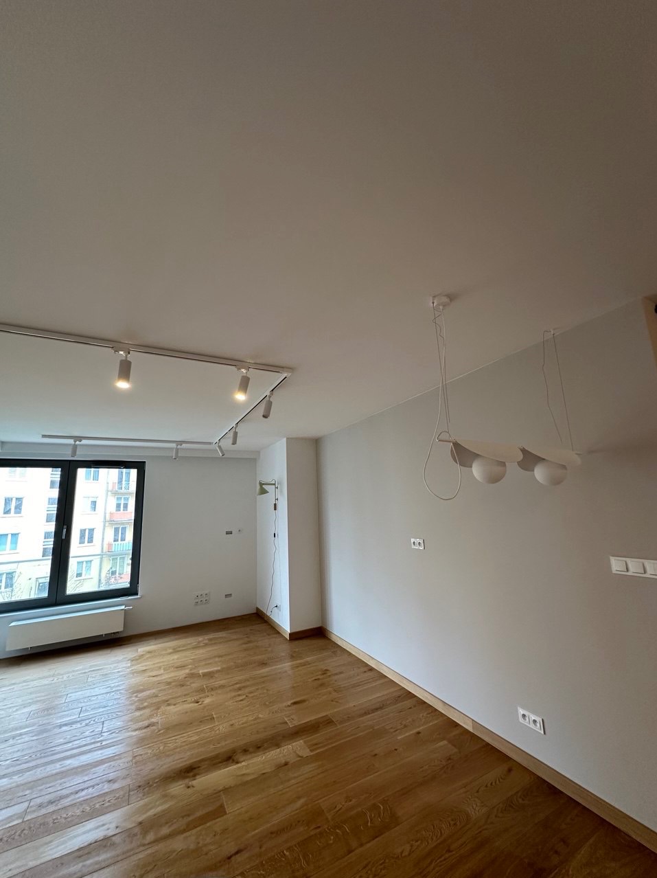 Pusty pokój po remoncie z parkietem w odcieniach brązu, białymi ścianami z widocznymi gniazdkami elektrycznymi, nowoczesnym oświetleniem sufitowym i dużym oknem z widokiem na budynek mieszkalny.