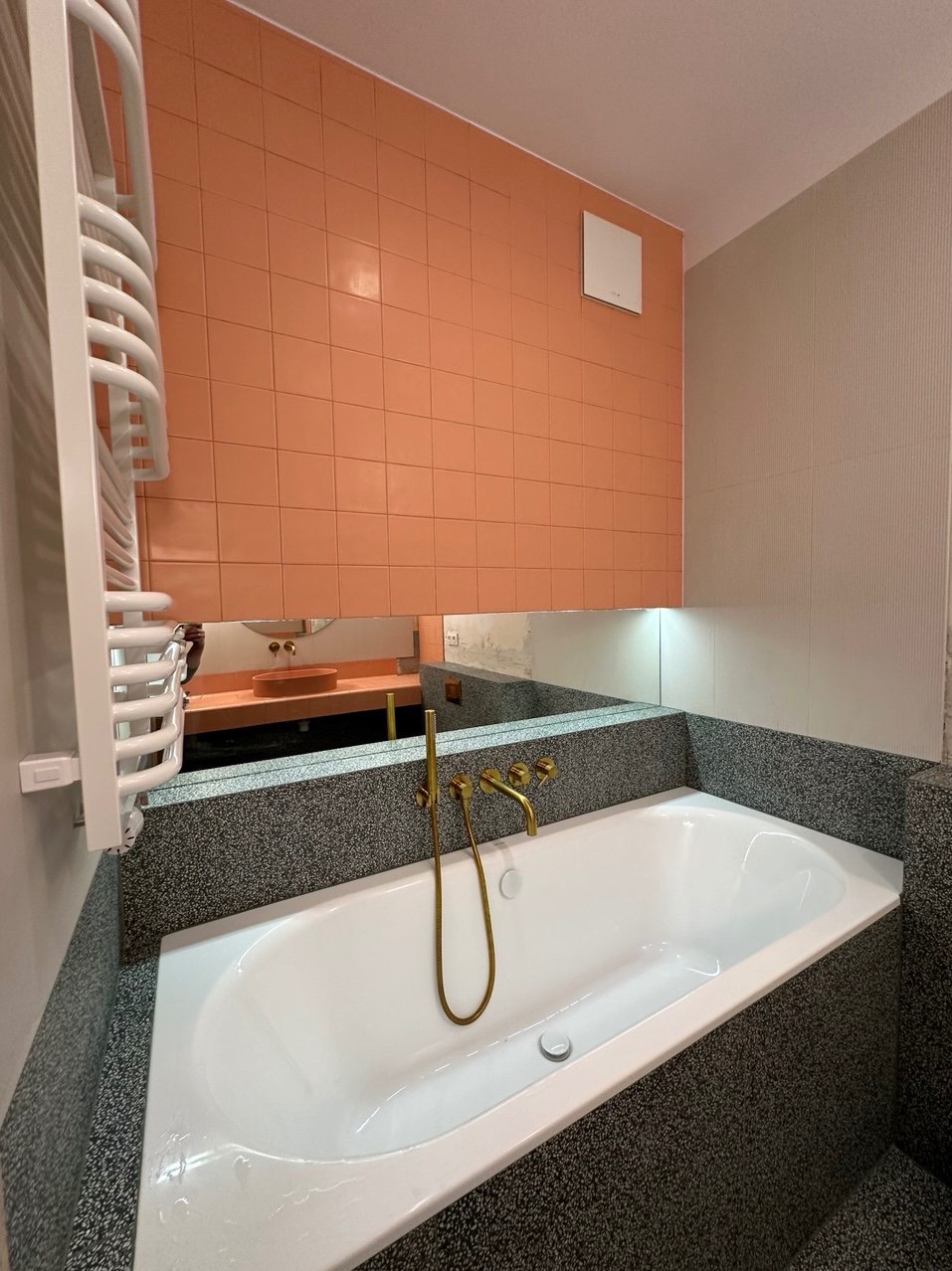 Wykończona łazienka z białą wanną obudowaną szarym lastryko, złotą armaturą i pomarańczowymi płytkami na ścianie, widoczna również w lustrze toaletka z umywalką.