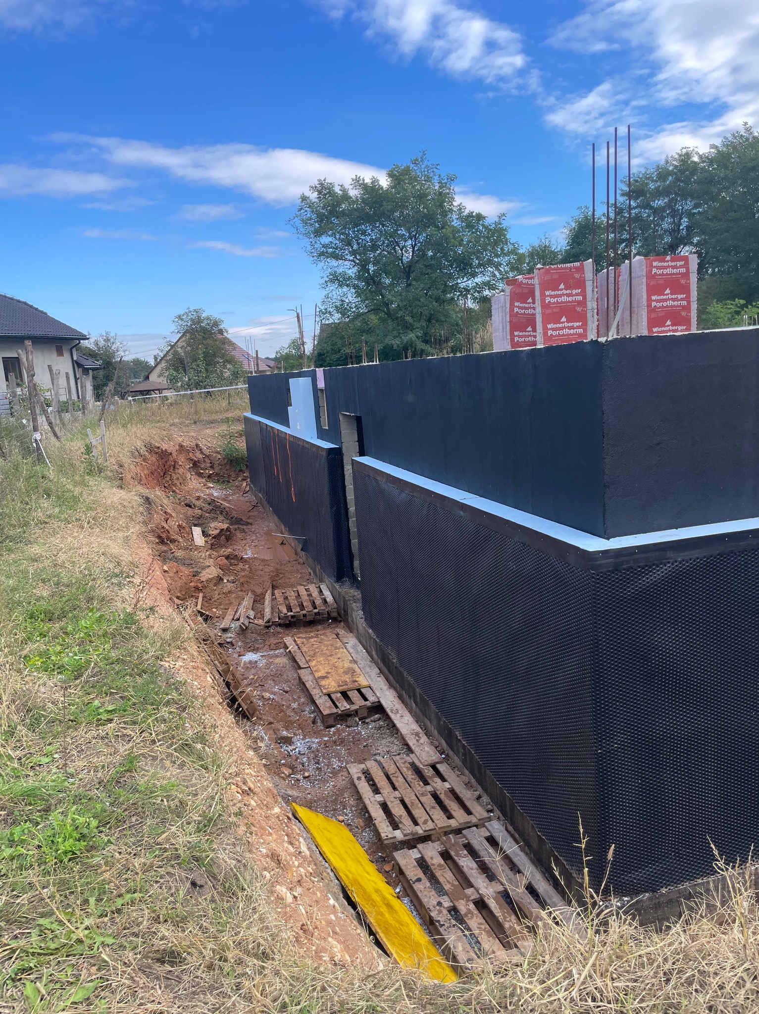 Fundament domu w trakcie budowy, zabezpieczony czarną izolacją przeciwwilgociową, z widocznymi paletami i bloczkami Wienerberger Porotherm, częściowo zasypany ziemią.