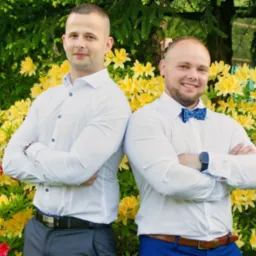 Dwóch mężczyzn w białych koszulach stoi plecami do siebie na tle kwitnącego krzewu z żółtymi i czerwonymi kwiatami, jeden ma niebieską muchę, drugi szare spodnie.