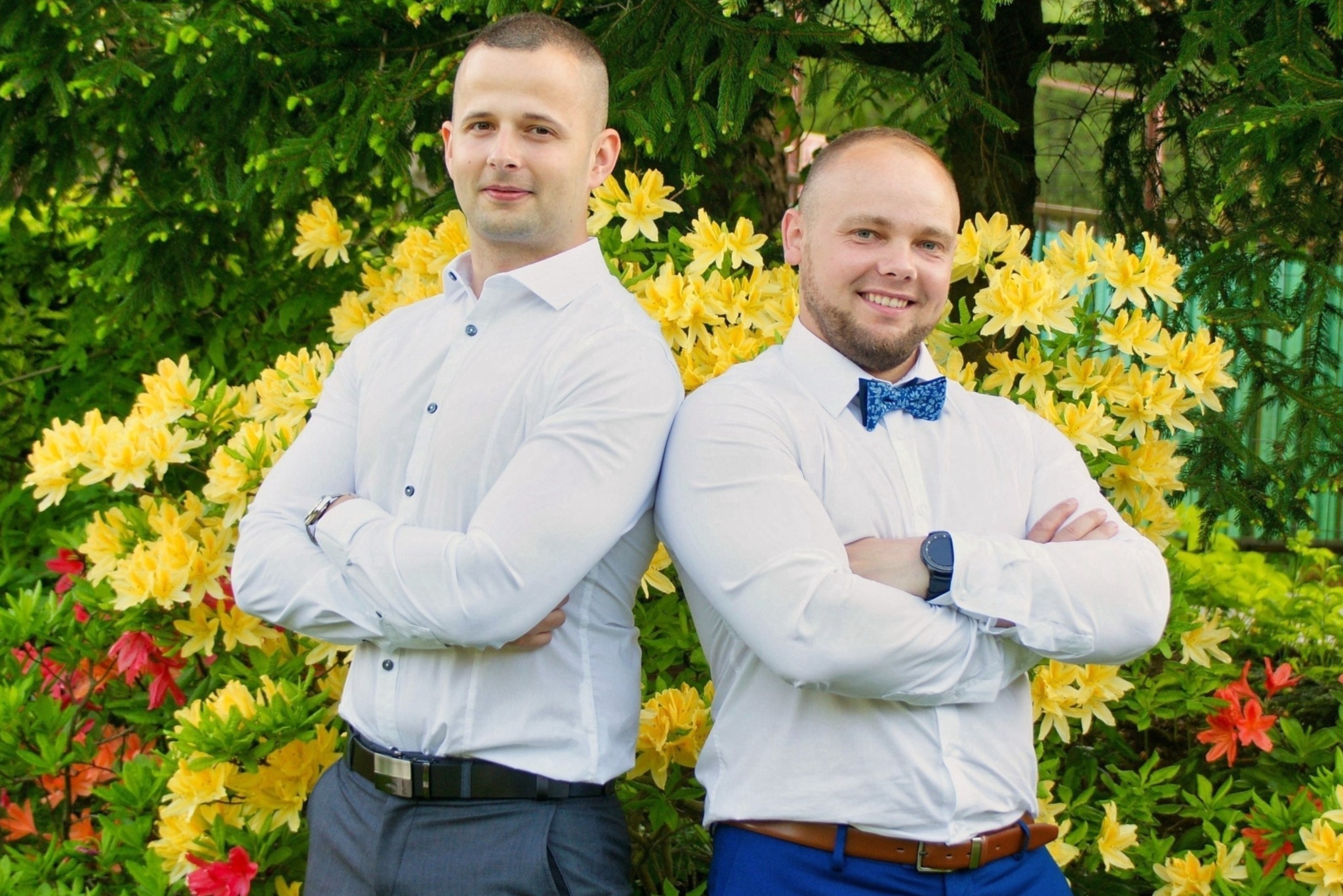 Dwóch mężczyzn w białych koszulach stoi plecami do siebie na tle kwitnącego krzewu z żółtymi i czerwonymi kwiatami, jeden ma niebieską muchę, drugi szare spodnie.