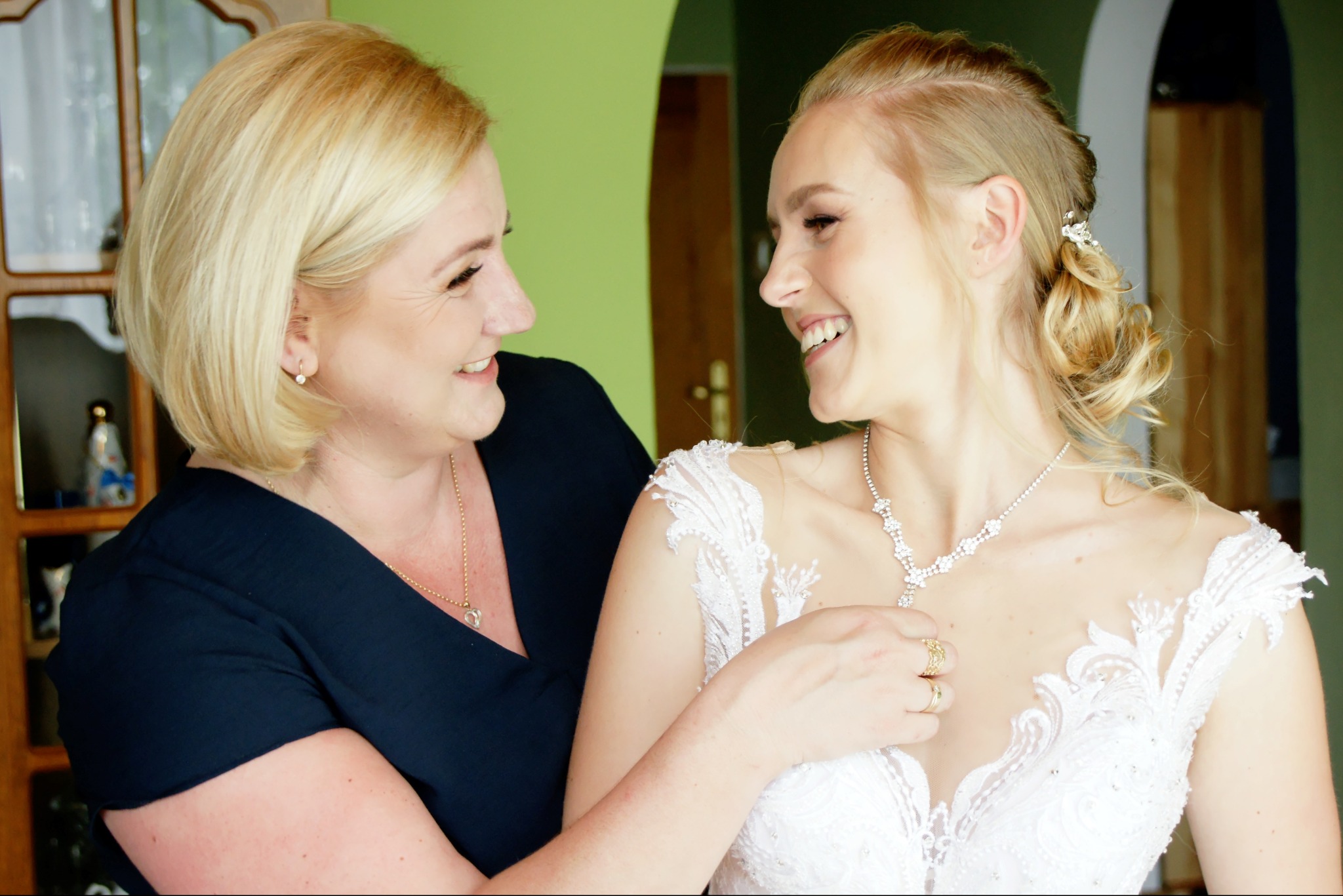 Panna młoda w białej sukni z koronkowymi zdobieniami uśmiecha się do kobiety w granatowej bluzce, która poprawia jej naszyjnik.