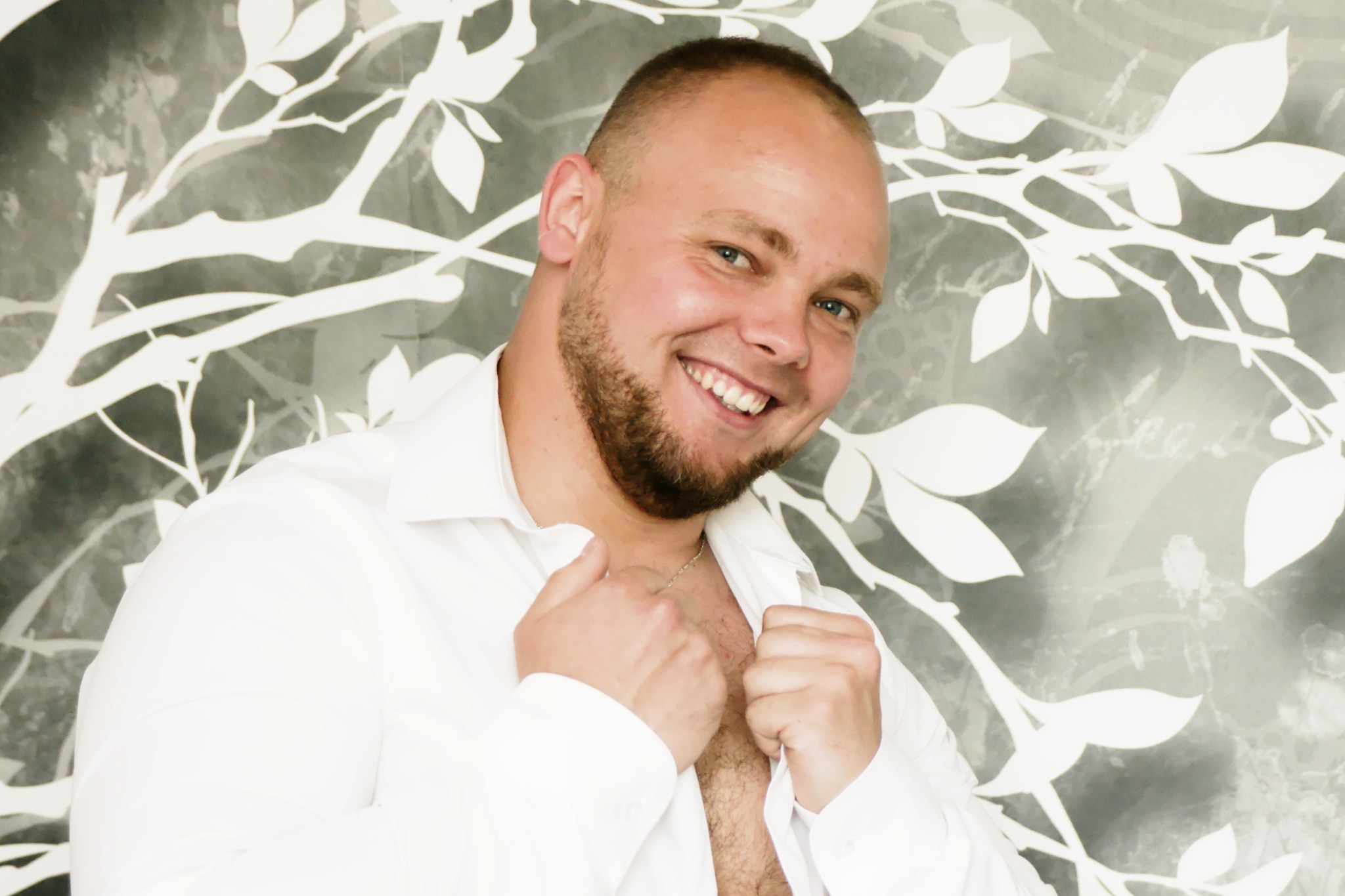 Uśmiechnięty mężczyzna z brodą w białej koszuli, pozujący na tle ściany z motywem białych gałęzi i liści.