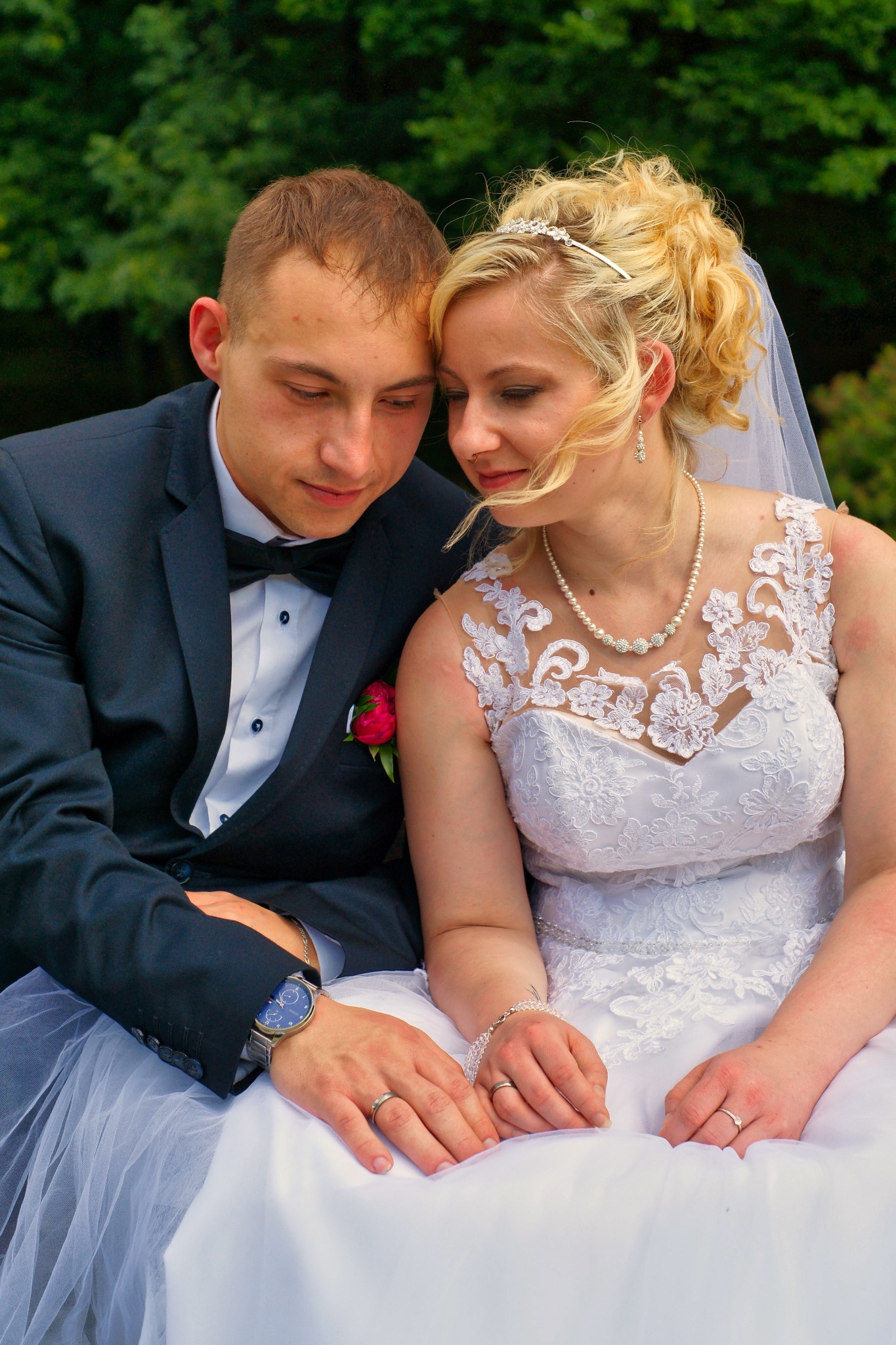 Para młoda: blondynka w koronkowej sukni i mężczyzna w granatowym smokingu z muszką, siedzą blisko siebie, patrząc w dół, dłonie spoczywają na białej sukni, w tle zielone drzewa.