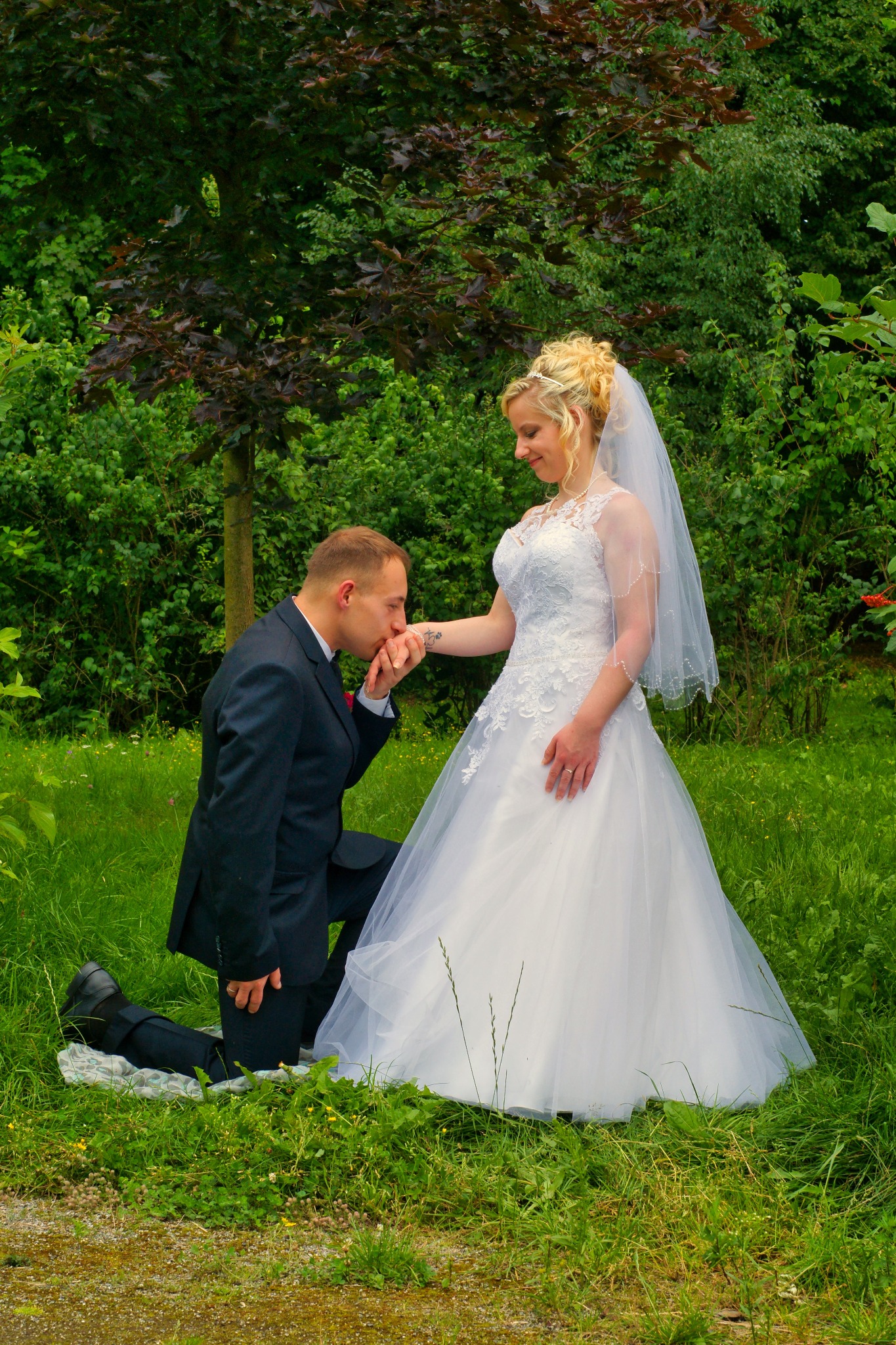 Pan młody w ciemnym garniturze klęczy na jednym kolanie, całując dłoń panny młodej w białej sukni ślubnej na tle zieleni drzew.