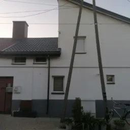 Dwukondygnacyjny dom z szarą dachówką, brązowymi drzwiami garażowymi i rowerem opartym o słup energetyczny