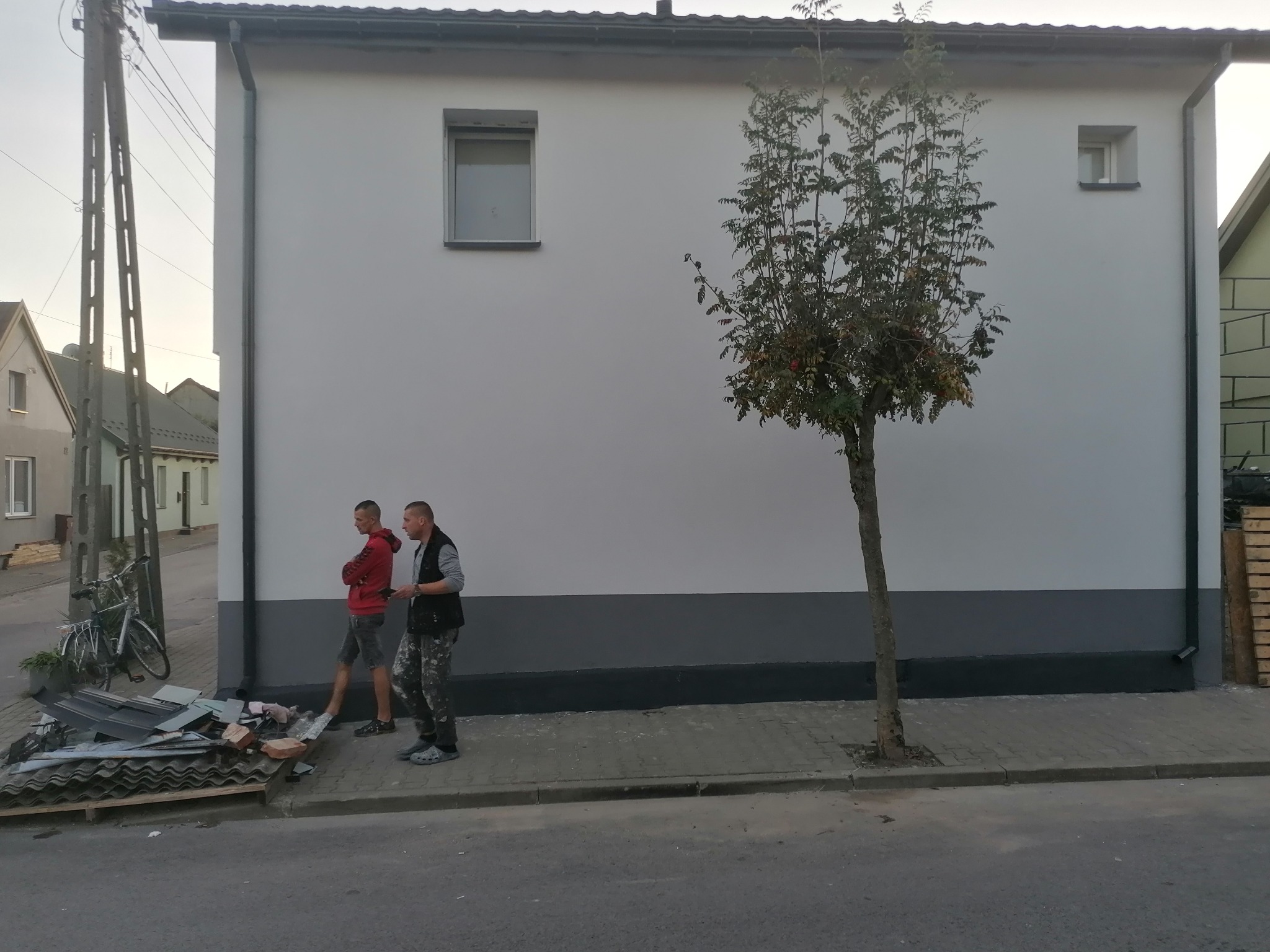 Dwukolorowa elewacja budynku z dwoma małymi oknami, świeżo pomalowana szarą farbą w dolnej części, z dwoma mężczyznami przechodzącymi obok i stertą gruzu budowlanego na chodniku.
