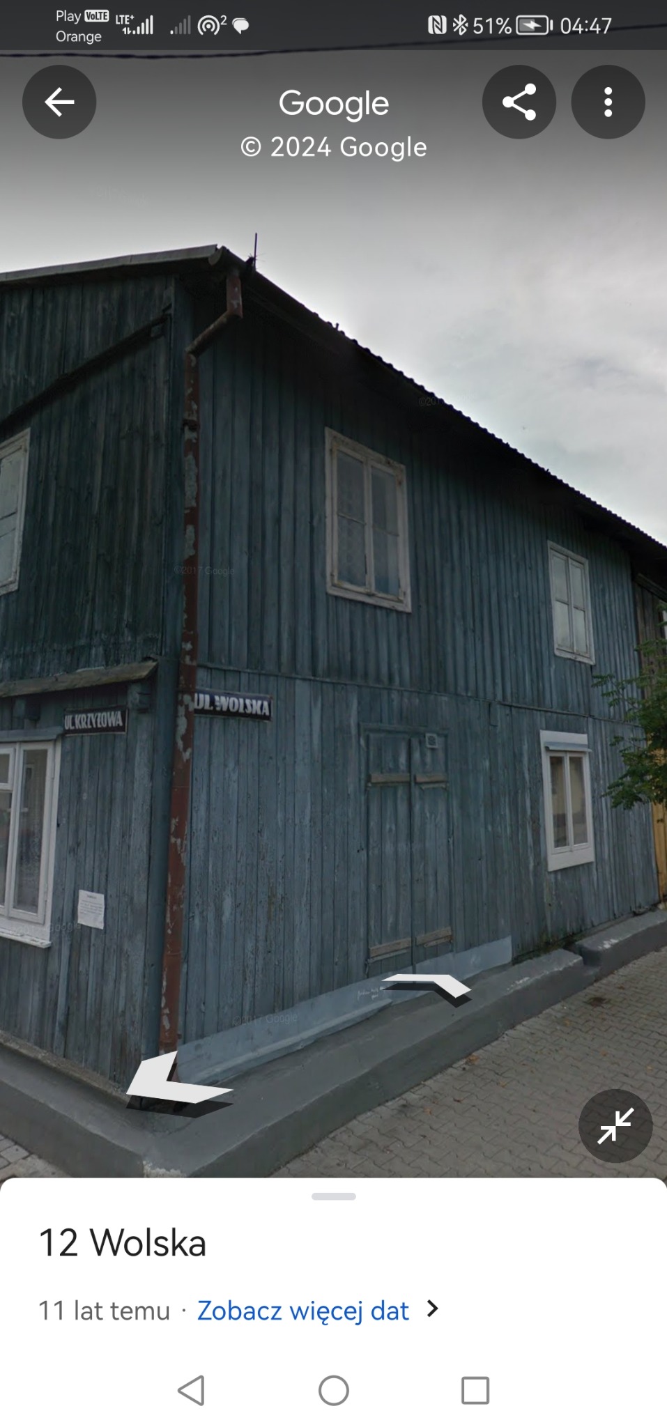 Dwukondygnacyjny, drewniany dom z szarej boazerii z białymi ramami okiennymi, widoczny z perspektywy ulicy z tabliczkami z nazwami ulic 'Ul. Wolska' i 'Ul. Krzyżowa'.