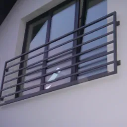 Prosta, metalowa barierka okienna z poziomymi szczeblami, zamontowana na jasnej elewacji budynku, widoczna z boku.