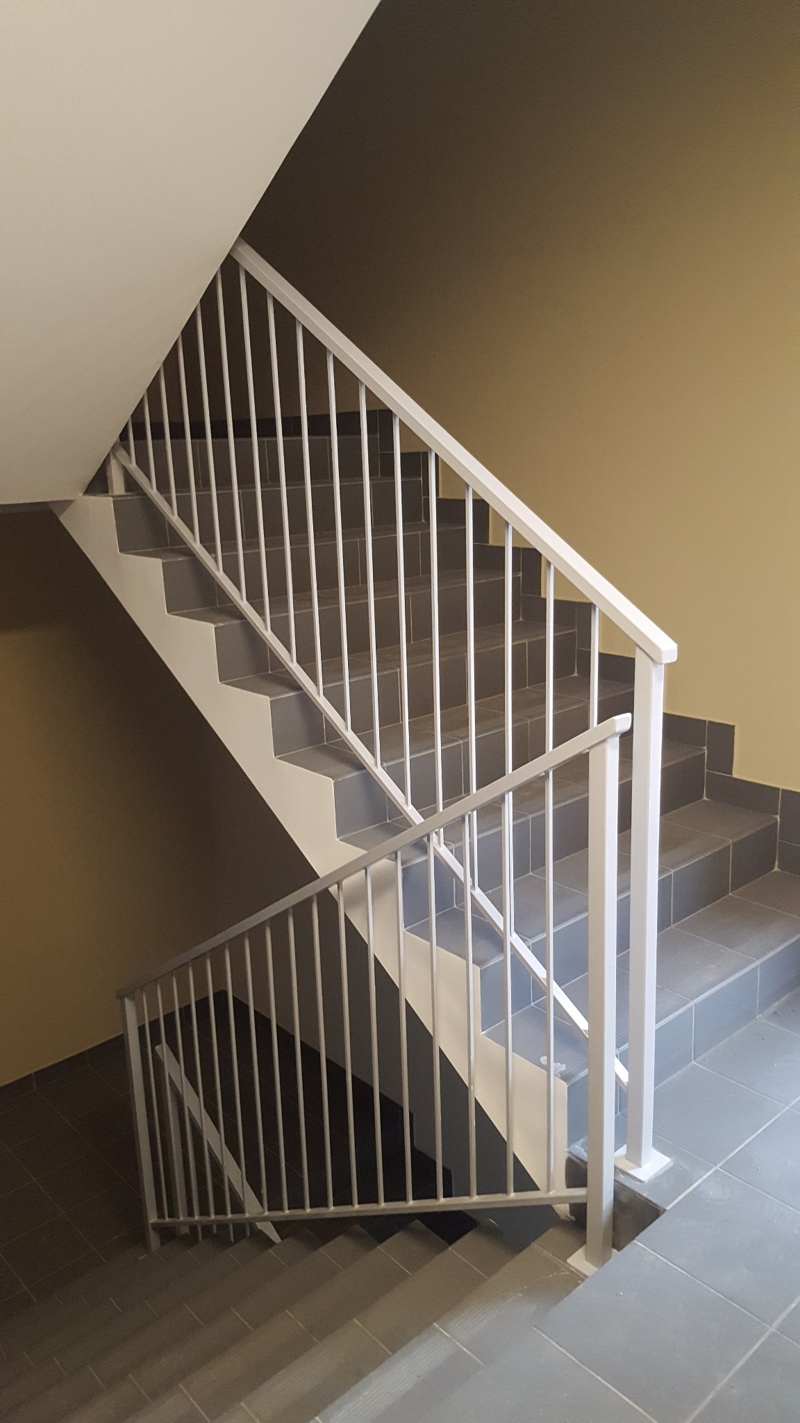 Klatka schodowa z szarymi płytkami i białą, metalową balustradą z pionowymi szczeblami, widok z dołu na górę.