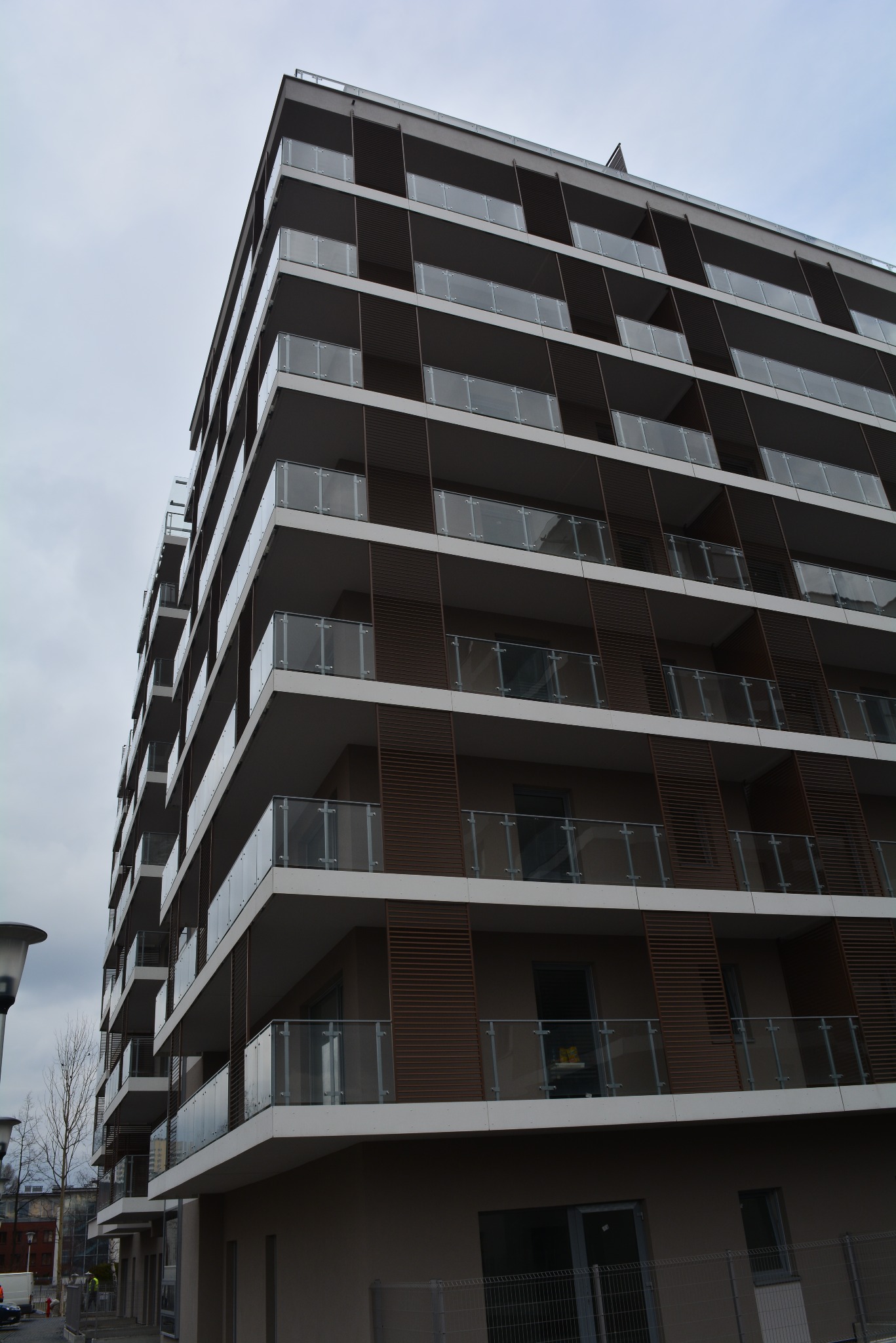 Wielopiętrowy budynek mieszkalny w Katowicach z balkonami zabezpieczonymi balustradami szklanymi, widoczne brązowe panele dekoracyjne między balkonami.