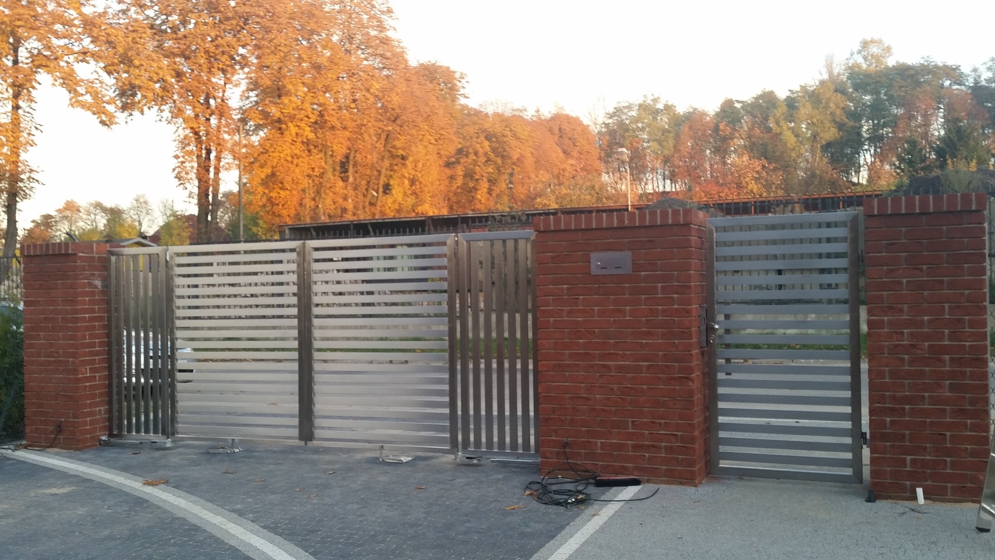 Automatyczna brama wjazdowa z poziomymi panelami i murowanymi słupkami z czerwonej cegły, widoczne przewody zasilające automatykę