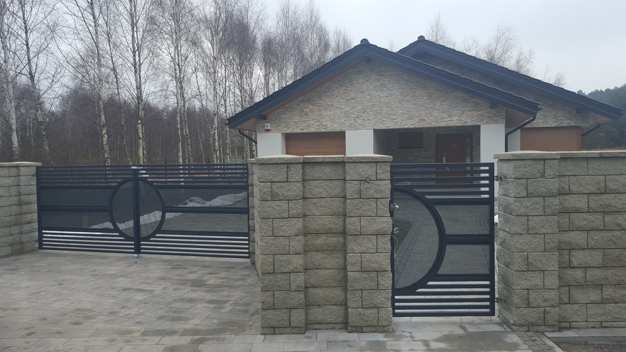 Nowoczesna, czarna brama wjazdowa z ażurowym, okrągłym wypełnieniem i kamiennymi słupkami, prowadząca do domu z elewacją z jasnego kamienia.