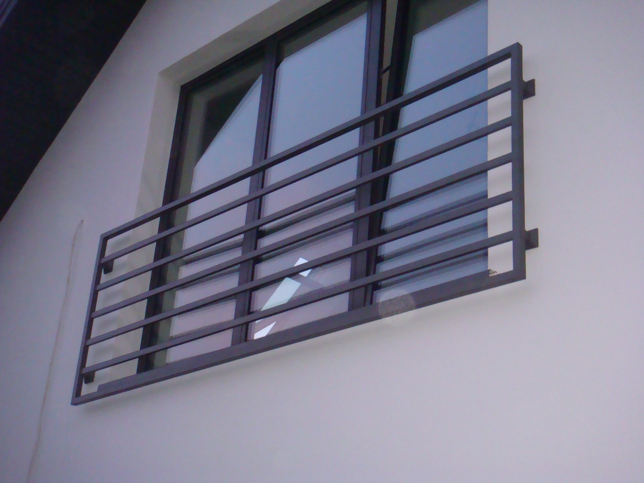 Prosta, metalowa barierka okienna z poziomymi szczeblami, zamontowana na jasnej elewacji budynku, widoczna z boku.