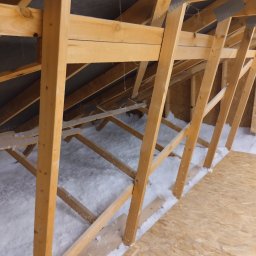 HC Izolacje - Poddasze w trakcie ocieplania: drewniana konstrukcja więźby dachowej wypełniona białą wełną mineralną, widoczna płyta OSB na podłodze, szara membrana dachowa w tle.