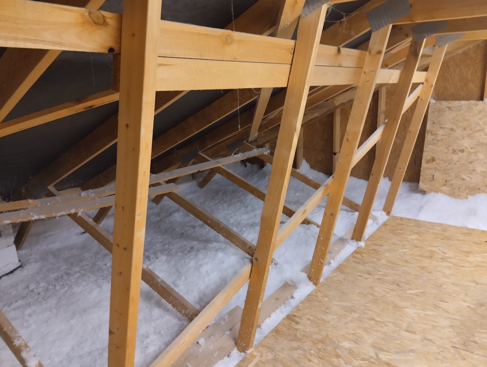 Poddasze w trakcie ocieplania: drewniana konstrukcja więźby dachowej wypełniona białą wełną mineralną, widoczna płyta OSB na podłodze, szara membrana dachowa w tle.