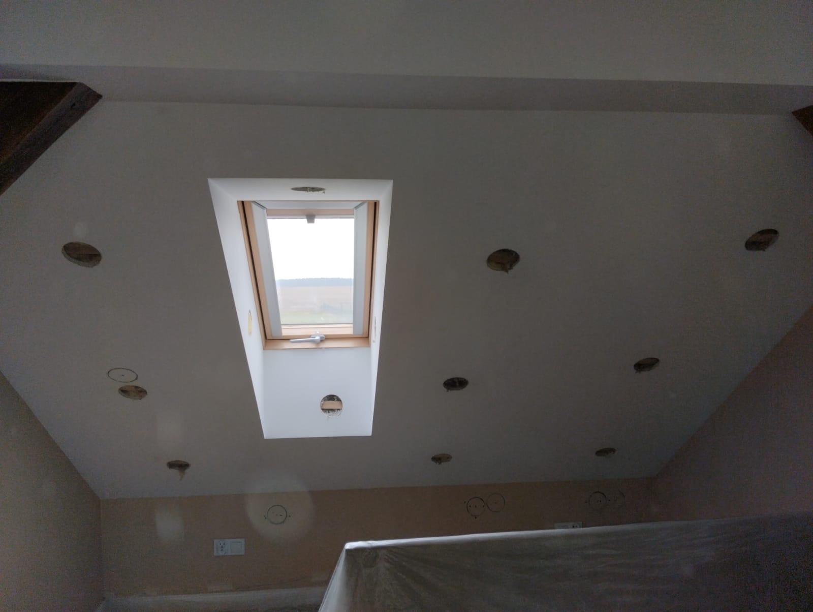Poddasze po ociepleniu z zamontowanym oknem dachowym i wywierconymi otworami pod oświetlenie punktowe. Widoczny fragment drewnianej belki stropowej.
