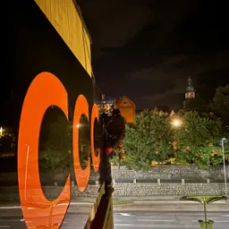 Pracownik w uprzęży, stojący na krawędzi budynku podczas nocnych prac na wysokości, z widocznym dużym, pomarańczowym logo na elewacji, w tle panorama miasta z wieżą kościelną.