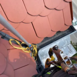 Widok z góry na czerwony dach z dachówki ceramicznej, rynnę z osadzonymi zanieczyszczeniami oraz sprzęt asekuracyjny i narzędzie do czyszczenia, sugerujący prace na wysokości.