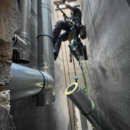 Usługi alpinistyczne Oskar Foitzik - Pracownik na linach montuje rury wentylacyjne w wąskim szybie, widoczne elementy instalacji i fragment okna.